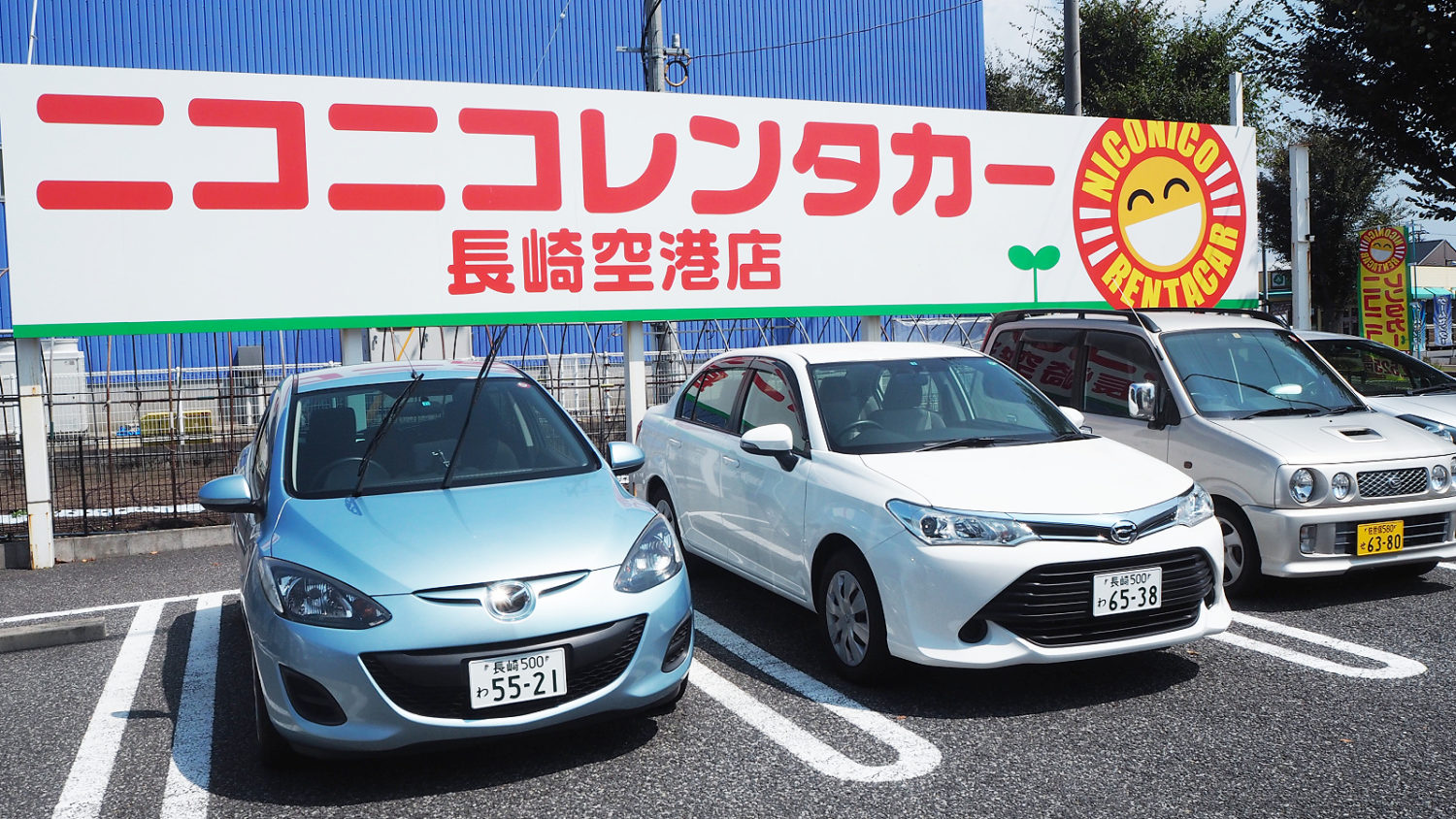 Blue, White, and Red Japanese cars under a NICONICO Rent a Car billboard.